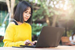 A female in glasses working on laptop outside at a table.